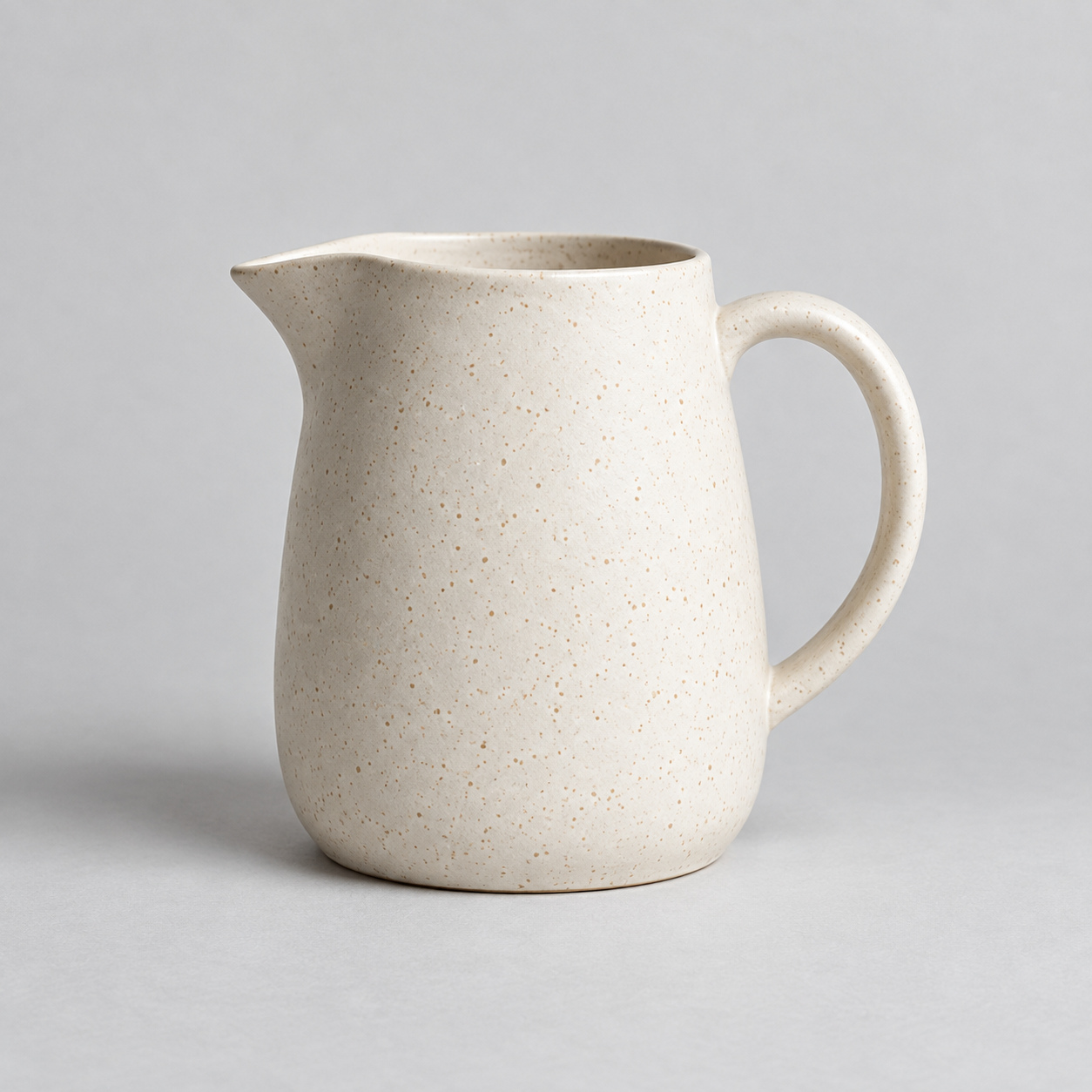 Speckled ivory ceramic pitcher with a curved handle on a gray studio surface